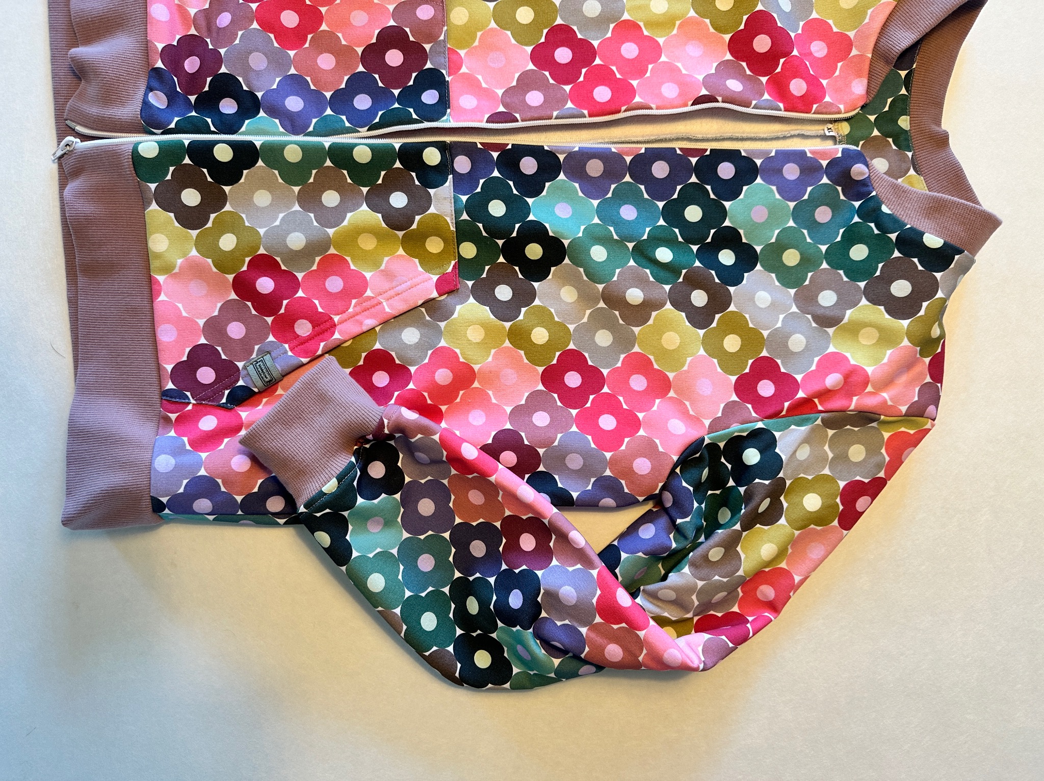 Rozłożona bluza dziecięca z kapturem, uszyta z kolorowej dzianiny w kwiatowy wzór, z widocznym zamkiem błyskawicznym i ściągaczami w kolorze lila.