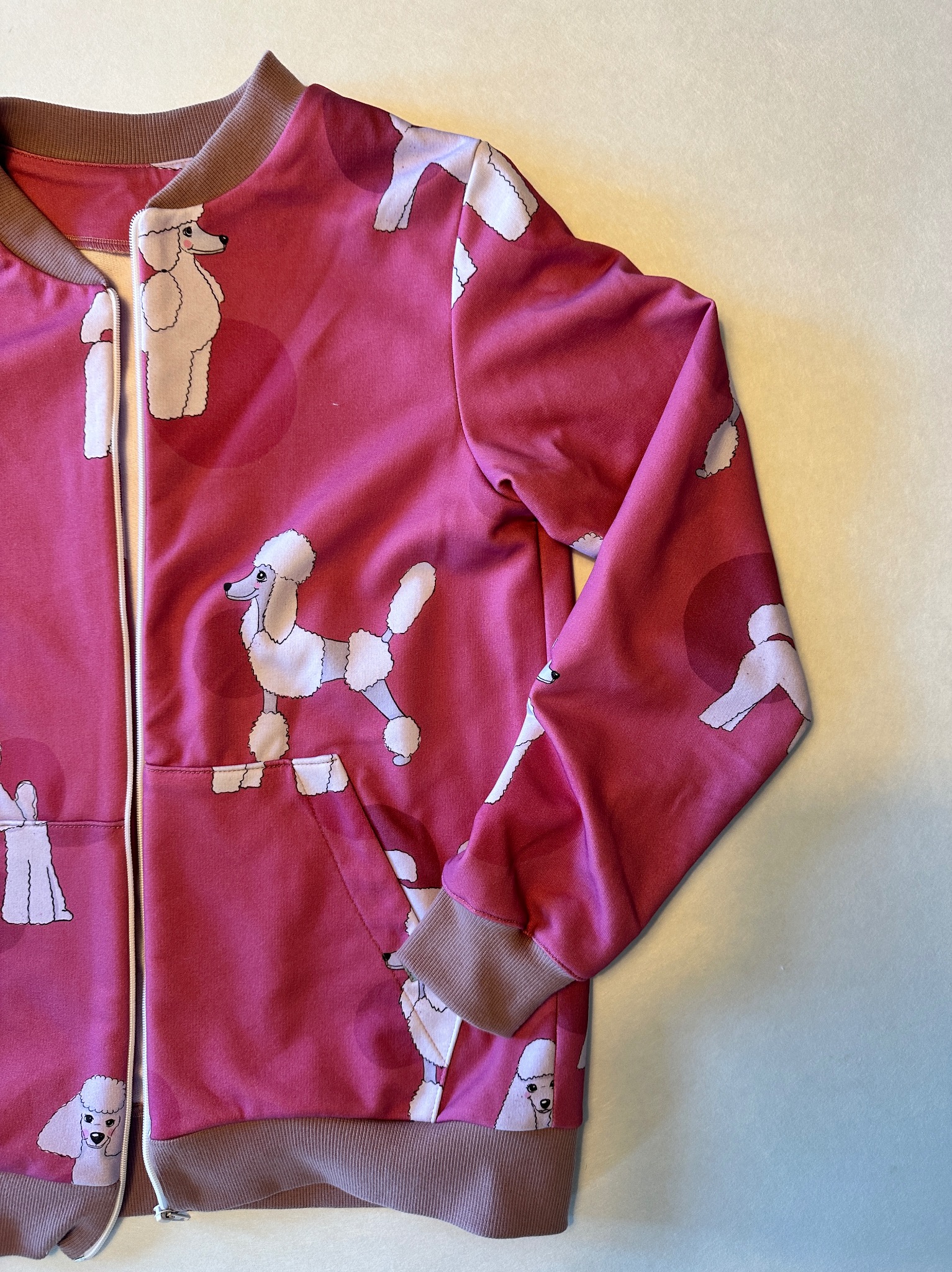 Rozpięta bluza bomberka w odcieniach różu z nadrukiem białych pudli, brązowe ściągacze przy mankietach, kołnierzu i na dole, leży na jasnym tle.