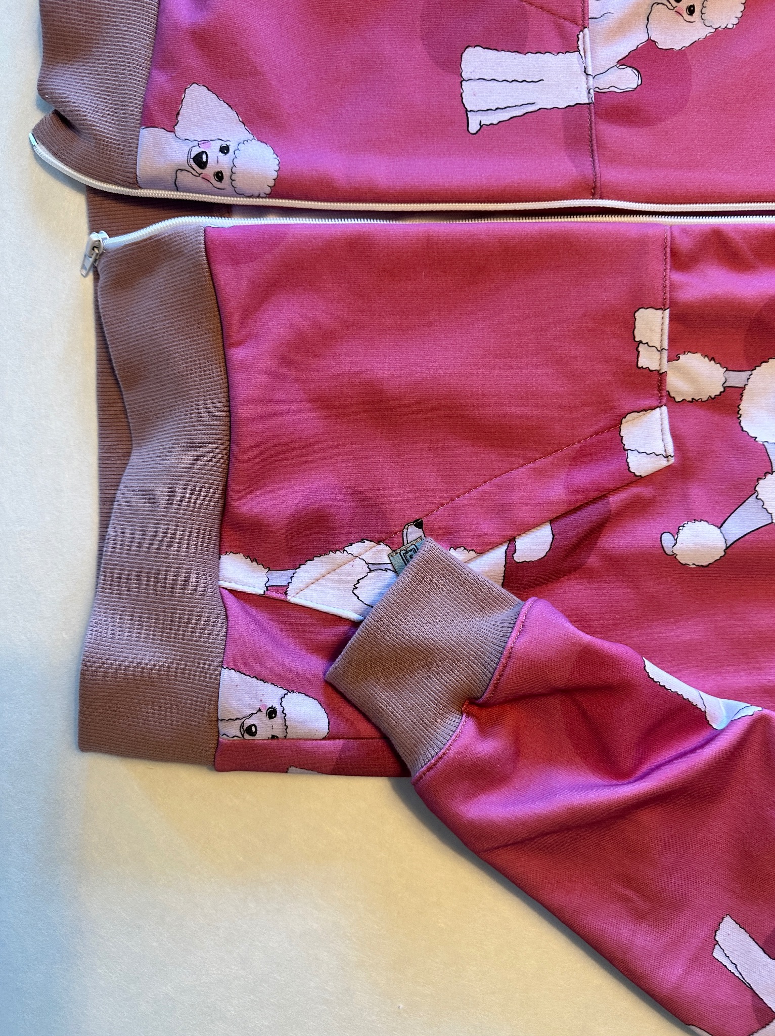 Różowa bluza dresowa rozpinana z nadrukiem białych pudli, mankiety i ściągacz w kolorze brudnego różu, widoczny zamek błyskawiczny i fragment kieszeni.