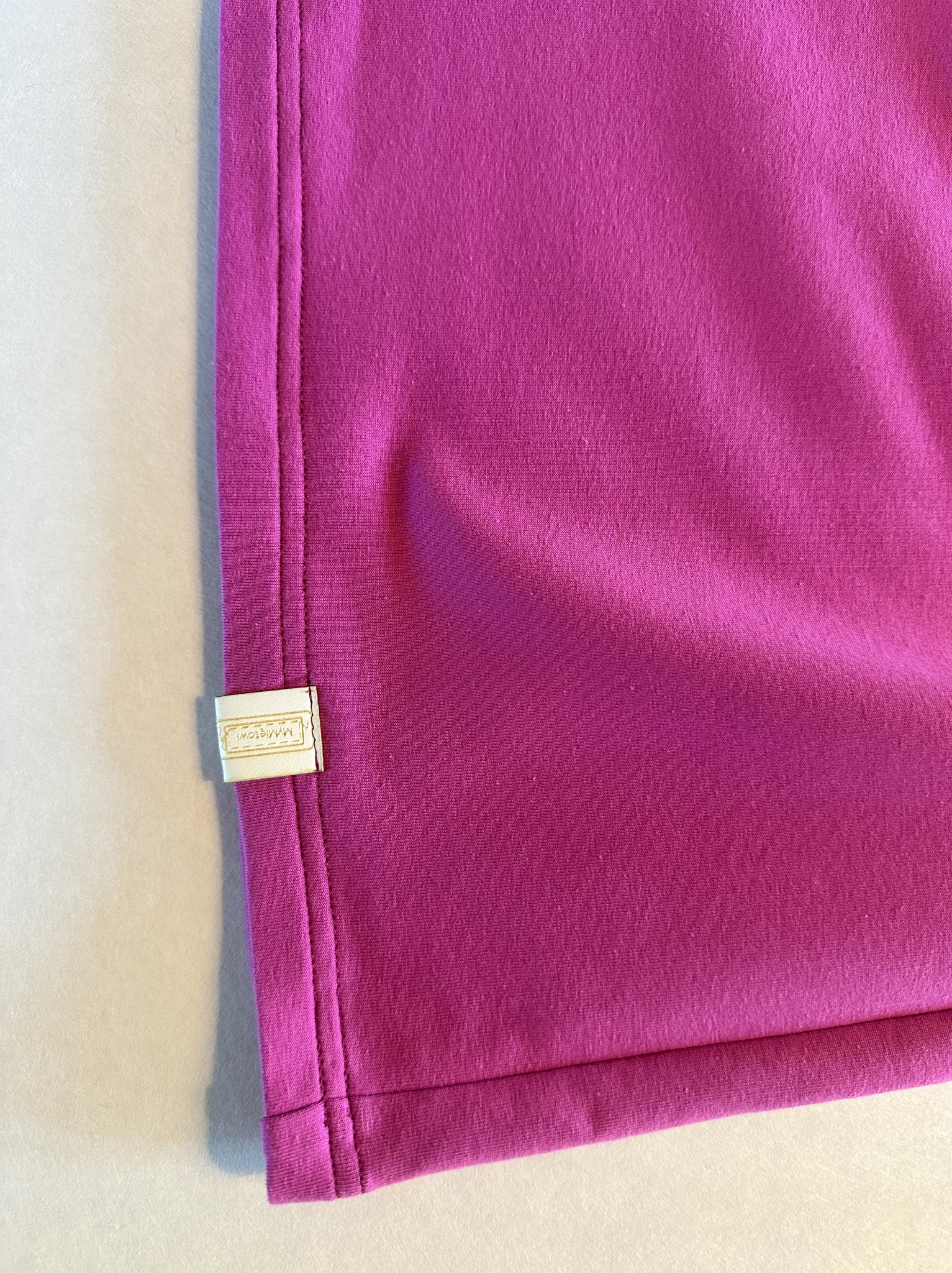 Fragment magenta tkaniny z widocznym szwem i wszytą metką z nazwą firmy, leżącej na jasnej powierzchni.