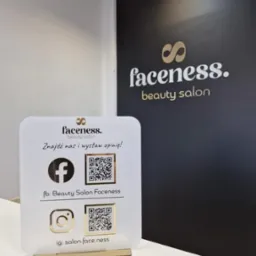 Tabliczka z informacjami o profilach w mediach społecznościowych salonu kosmetycznego Faceness, umieszczona na białym blacie, w tle czarna ściana z logo salonu.