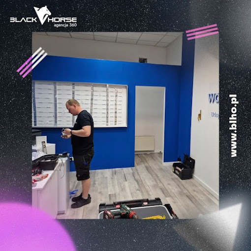 Pracownik w czarnej koszulce przygotowuje okleiny w nowo wykończonym biurze z niebieskimi ścianami i logo firmy na ścianie, widoczne narzędzia i skrzynki narzędziowe.