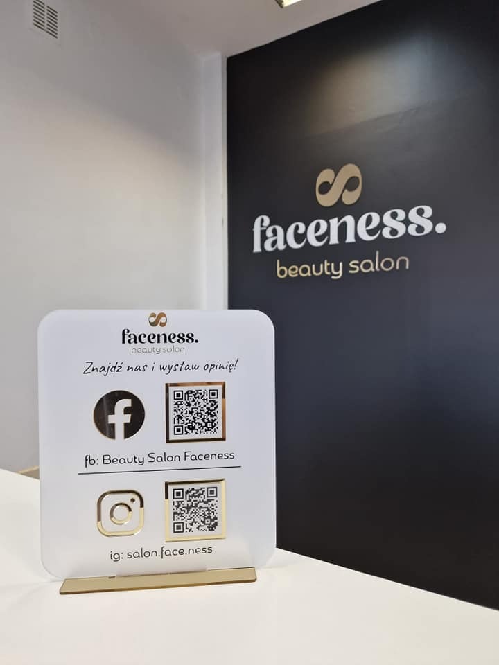 Tabliczka z informacjami o profilach w mediach społecznościowych salonu kosmetycznego Faceness, umieszczona na białym blacie, w tle czarna ściana z logo salonu.