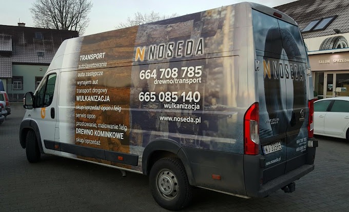 Biały samochód dostawczy z oklejeniem reklamowym firmy Noseda, oferującej transport, wulkanizację i sprzedaż drewna kominkowego, zaparkowany przed salonem sprzedaży.