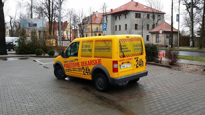 Żółty samochód dostawczy z napisem 'POGOTOWIE DEKARSKIE' i numerem telefonu, zaparkowany na brukowanym placu, w tle budynki mieszkalne i drzewa bez liści.