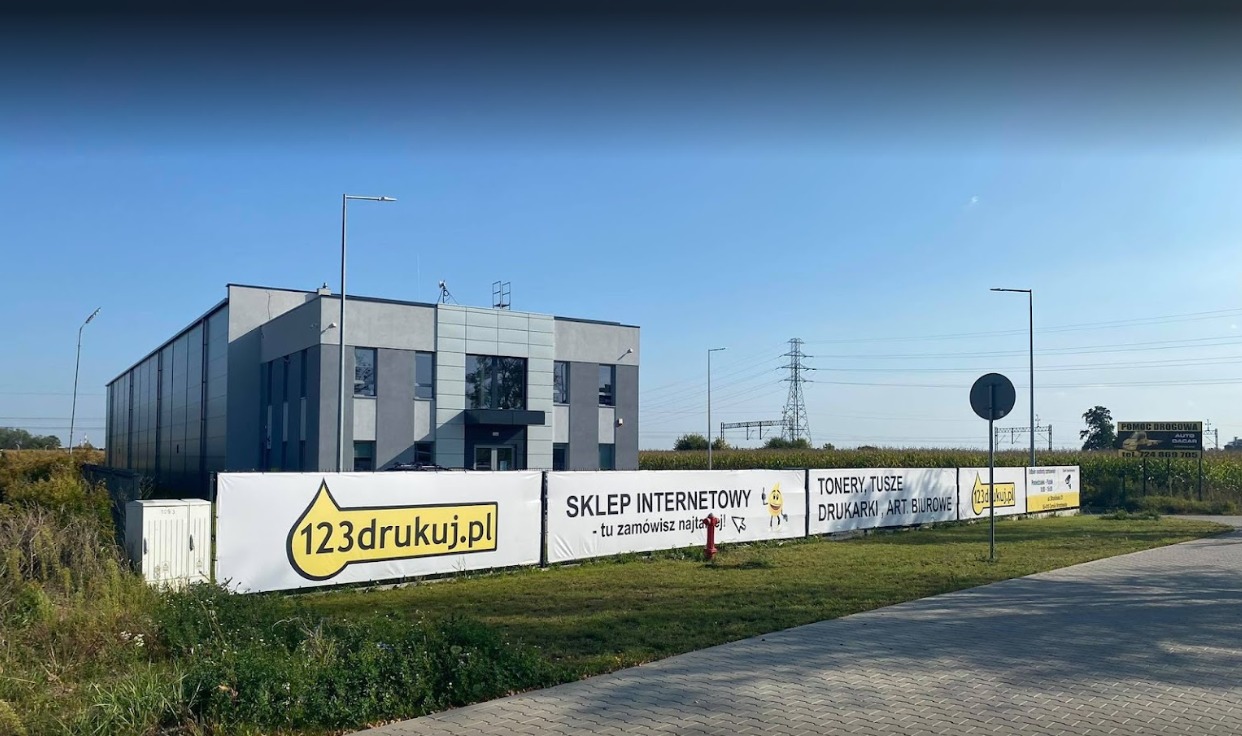 Banery reklamowe sklepu internetowego z artykułami biurowymi i drukarskimi przed budynkiem firmy, widoczny hydrant przeciwpożarowy i linia wysokiego napięcia w tle.