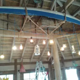 Pomysłowe oświetlenie wnętrza: odwrócona łódka z niebieskim pasem, zawieszona pod drewnianym sufitem na grubych linach, służy jako podstawa dla różnorodnych lamp i żarówek, tworząc unikalny...