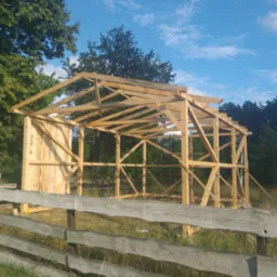Konstrukcja szkieletowa budynku w trakcie budowy, widoczna drewniana rama i częściowo obita ściana, na pierwszym planie drewniany płot, w tle drzewa i błękitne niebo z chmurami.