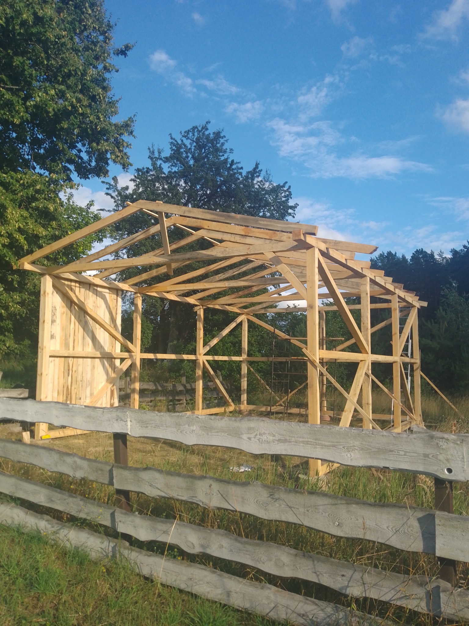 Konstrukcja szkieletowa budynku w trakcie budowy, widoczna drewniana rama i częściowo obita ściana, na pierwszym planie drewniany płot, w tle drzewa i błękitne niebo z chmurami.