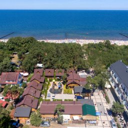 EMI DOM S.C SYLWIA MIŁOSZ STOSIO - Widok z lotu ptaka na kompleks drewnianych domków letniskowych z placem zabaw, położonych blisko piaszczystej plaży z tłumem ludzi i błękitnego morza.