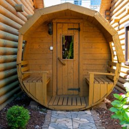 EMI DOM S.C SYLWIA MIŁOSZ STOSIO - Drewniana sauna w kształcie beczki z małym oknem i ławeczkami po bokach, umieszczona na zewnątrz przy ścianie z bali, prowadzi do niej ścieżka z kamiennej kostki.