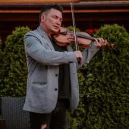 Skrzypek w szarej marynarce i podartych dżinsach gra na skrzypcach na tle zielonych krzewów i budynku z drewnianą fasadą.