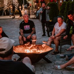 EMI DOM S.C SYLWIA MIŁOSZ STOSIO - Grupa osób relaksuje się przy ognisku, piekąc kiełbaski; w tle skrzypek gra na instrumencie, oświetlenie wieczorne.