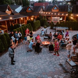 EMI DOM S.C SYLWIA MIŁOSZ STOSIO - Zorganizowana impreza plenerowa z ogniskiem, muzyką graną na skrzypcach, stołem do piłkarzyków i koszem do koszykówki, oświetlona girlandami żarówek, otoczona tujami i drewnianą zabudową.