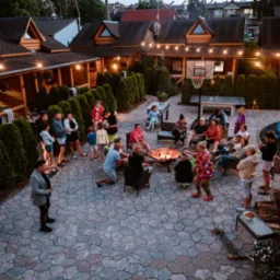 Zorganizowana impreza plenerowa z ogniskiem, muzyką graną na skrzypcach, stołem do piłkarzyków i koszem do koszykówki, oświetlona girlandami żarówek, otoczona tujami i drewnianą zabudową.