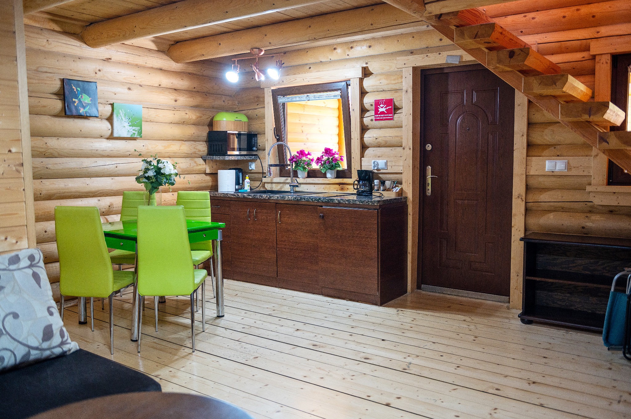 Wnętrze drewnianego domu z zielonym stołem i krzesłami, kwiatami w oknie, aneksem kuchennym i schodami.