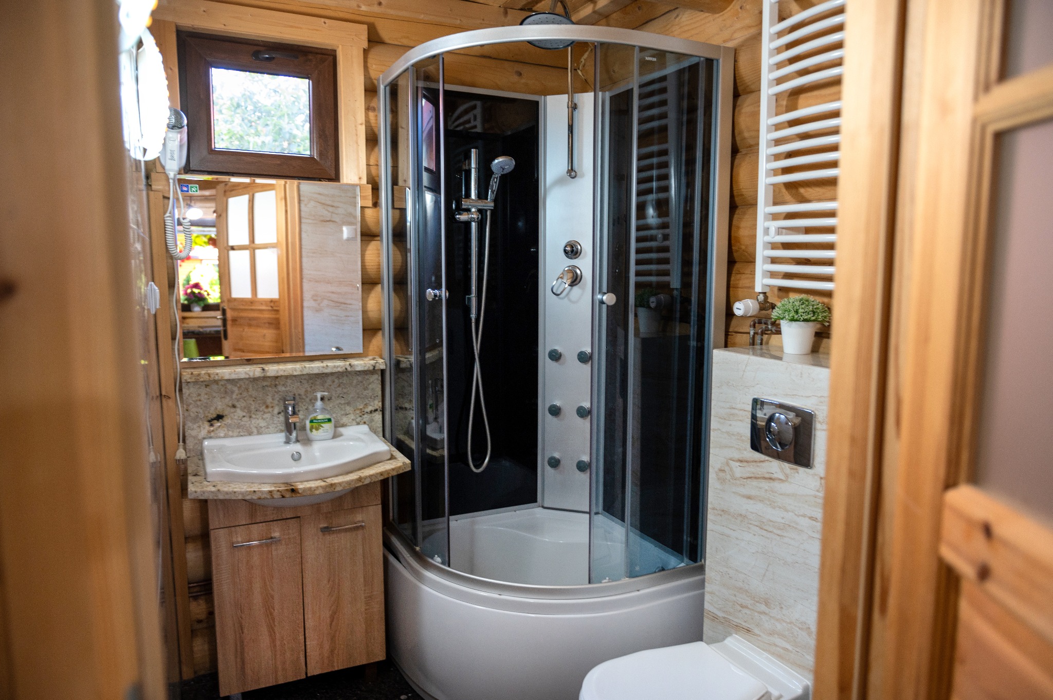 Wnętrze łazienki w drewnianym domku z kabiną prysznicową z hydromasażem, umywalką na szafce z kamiennym blatem, lustrem i grzejnikiem drabinkowym.