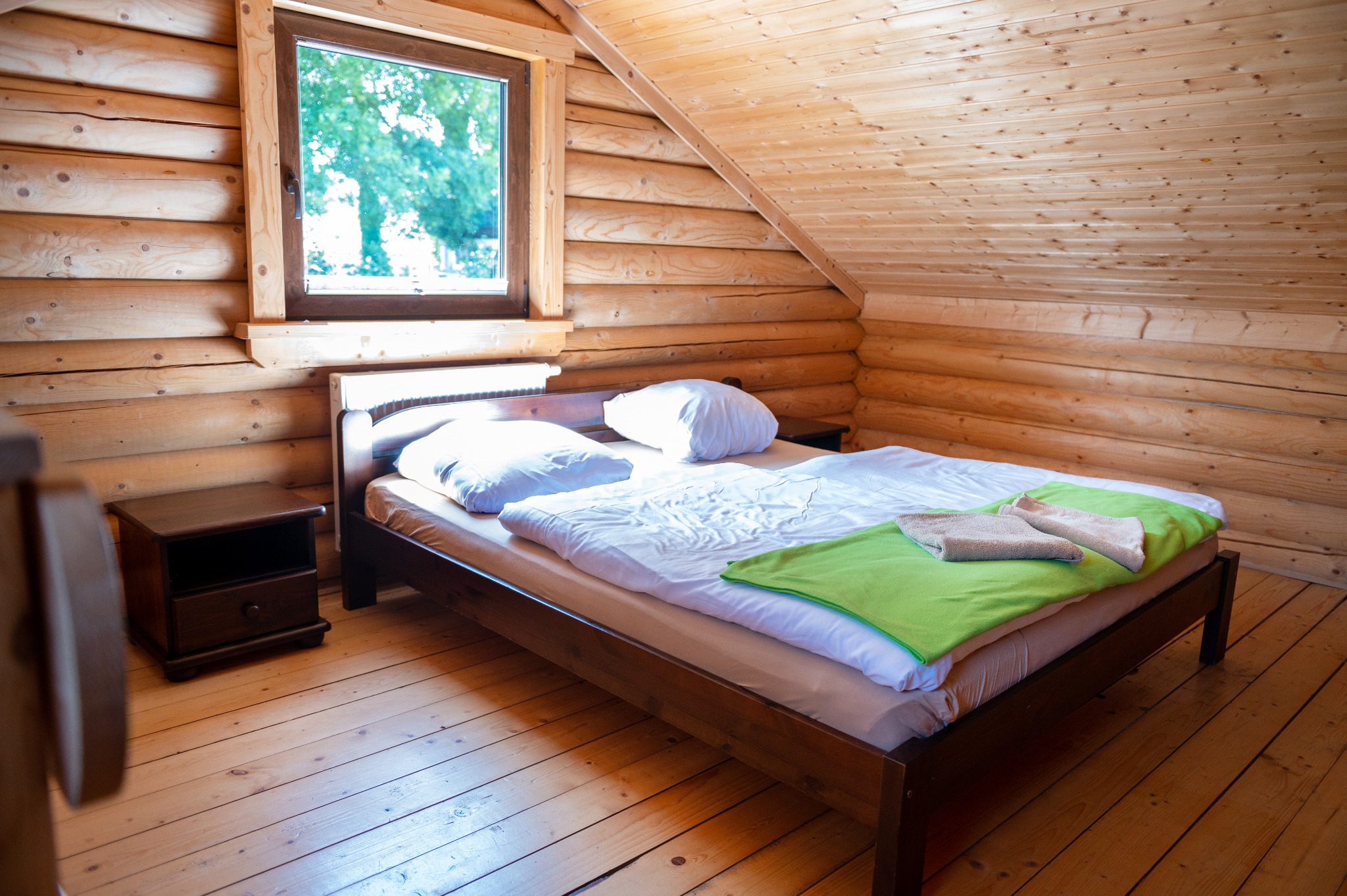 Drewniany pokój z łóżkiem, nakastlikiem i oknem, ściany i sufit z bali, pościel biała z zielonym akcentem i ręcznikami.