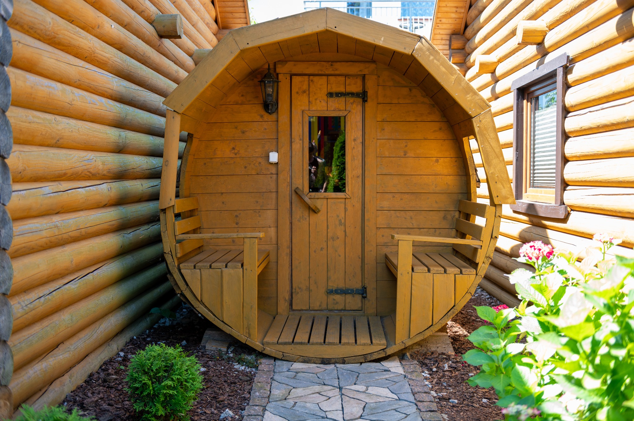 Drewniana sauna w kształcie beczki z małym oknem i ławeczkami po bokach, umieszczona na zewnątrz przy ścianie z bali, prowadzi do niej ścieżka z kamiennej kostki.