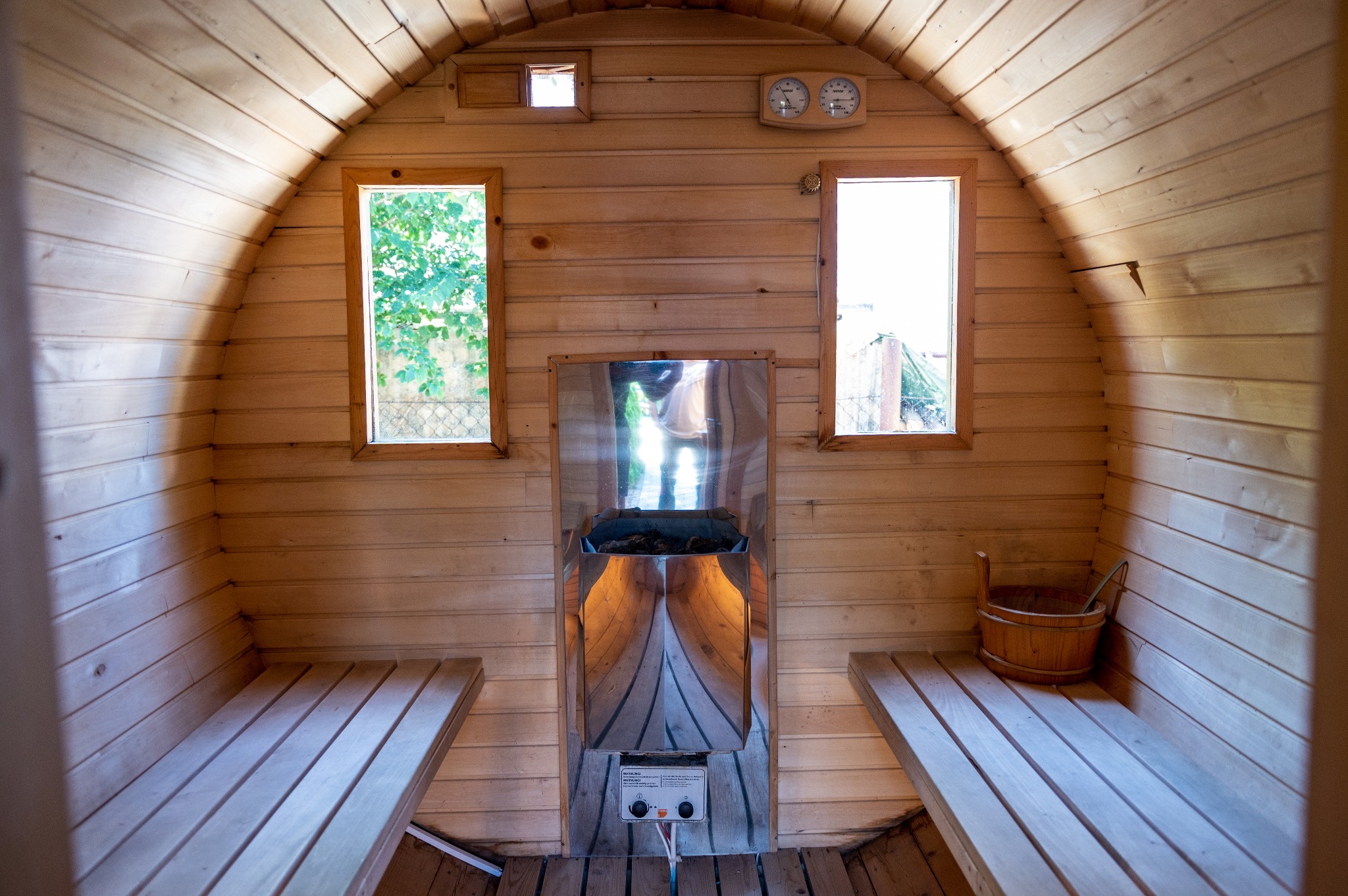 Wnętrze sauny beczki z piecem elektrycznym, drewnianymi ławkami i oknami z widokiem na zieleń, termometry na ścianie.