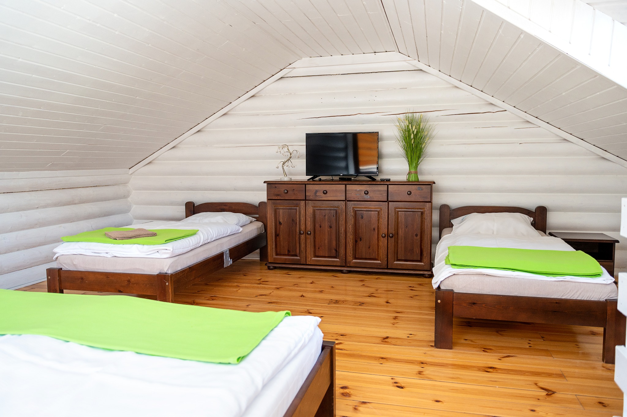 Pokój na poddaszu z trzema łóżkami pojedynczymi, drewnianą komodą z telewizorem i dekoracjami, białe ściany z bali i drewniana podłoga.