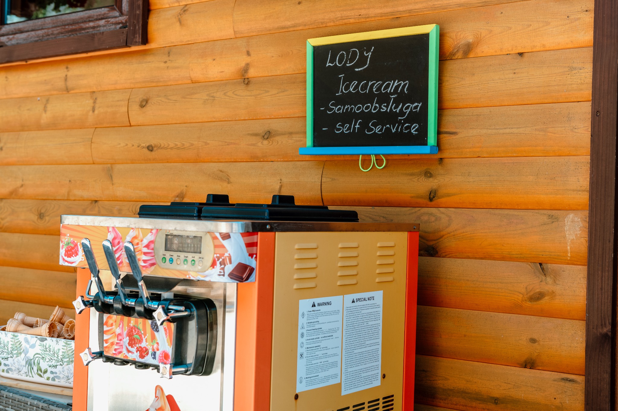 Automat do lodów samoobsługowych z tablicą informacyjną 'Lody - Icecream - Samoobsługa - self service' na tle drewnianej ściany.