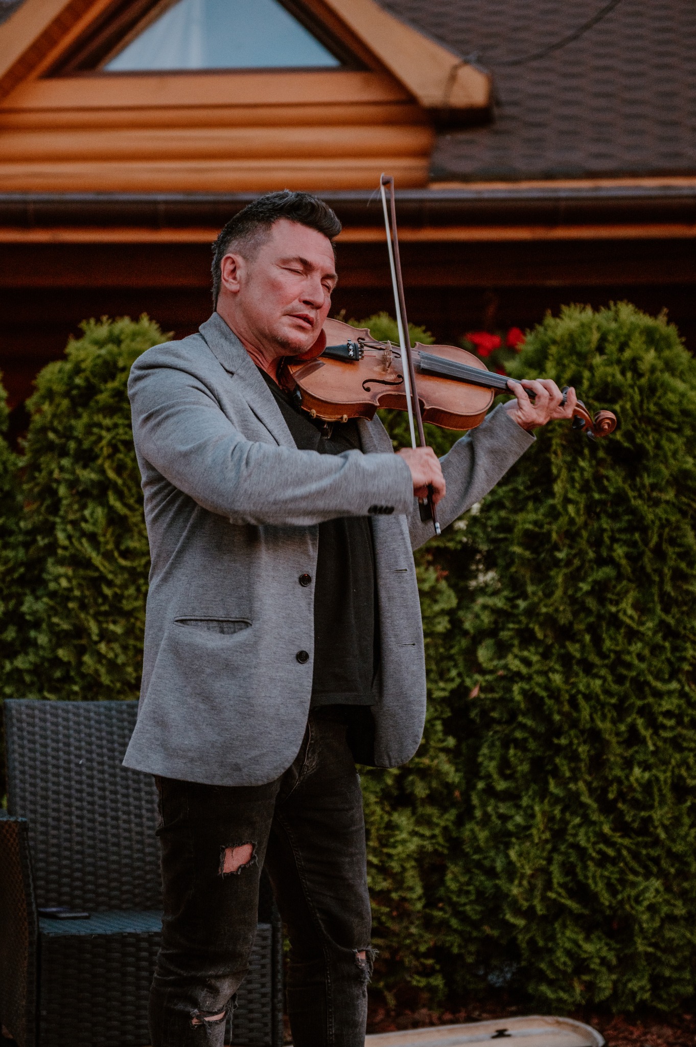 Skrzypek w szarej marynarce i podartych dżinsach gra na skrzypcach na tle zielonych krzewów i budynku z drewnianą fasadą.