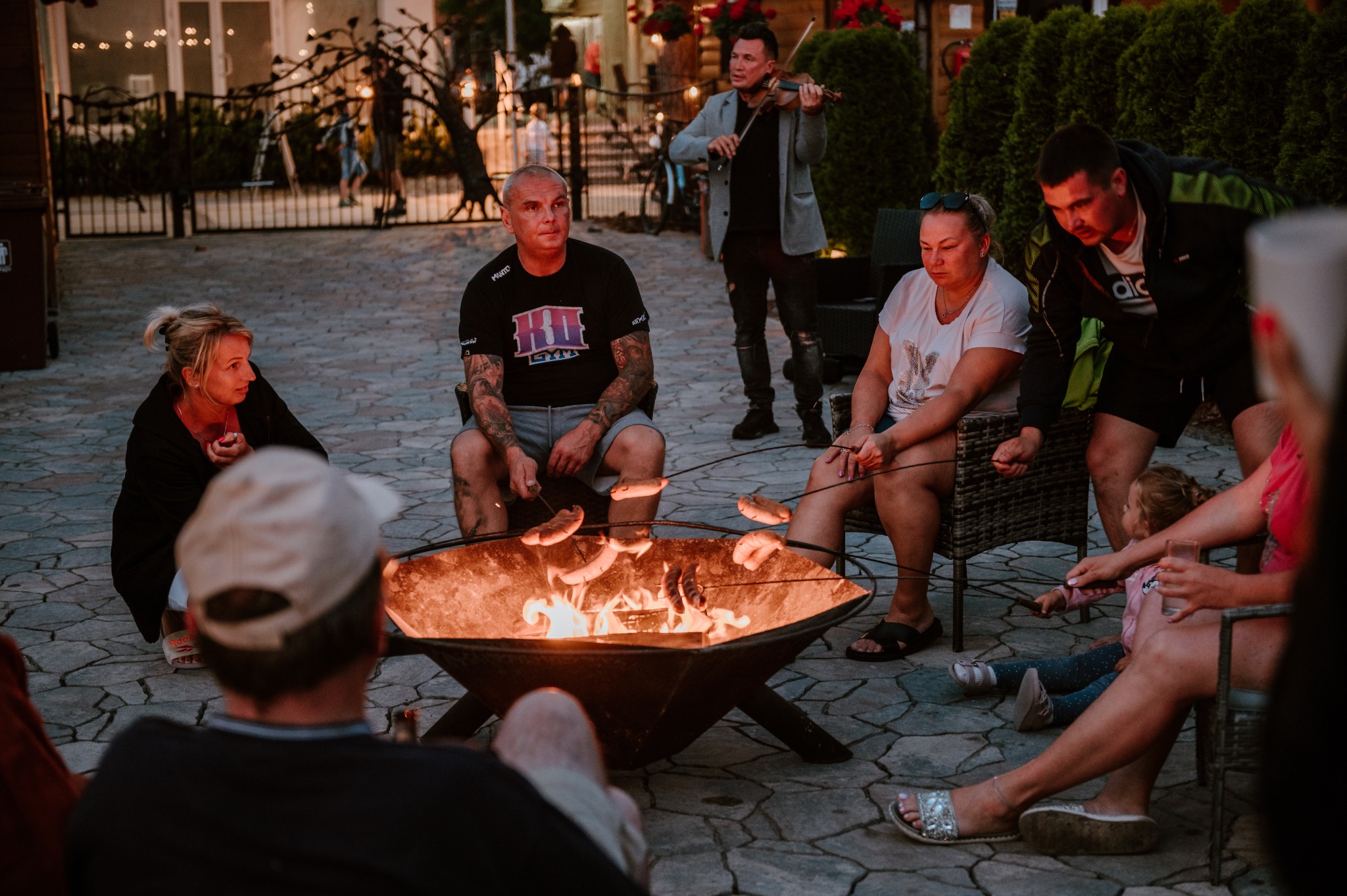 Grupa osób relaksuje się przy ognisku, piekąc kiełbaski; w tle skrzypek gra na instrumencie, oświetlenie wieczorne.