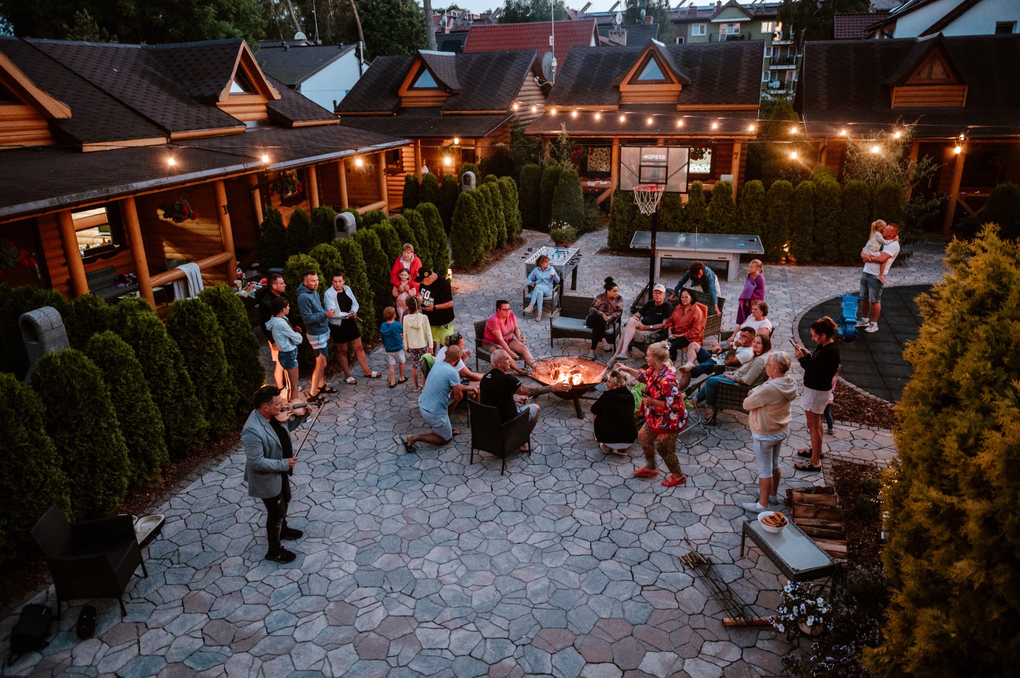 Zorganizowana impreza plenerowa z ogniskiem, muzyką graną na skrzypcach, stołem do piłkarzyków i koszem do koszykówki, oświetlona girlandami żarówek, otoczona tujami i drewnianą zabudową.
