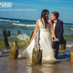 Sesja ślubna na plaży z drewnianymi palami wystającymi z wody, para młoda patrzy na siebie z uśmiechem, widoczne logo studia fotograficznego w lewym górnym rogu.