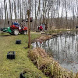 Prace porządkowe nad brzegiem stawu: traktor z kosiarką, ekipa usuwa gałęzie, czarne donice na brzegu, odbicie drzew w wodzie.