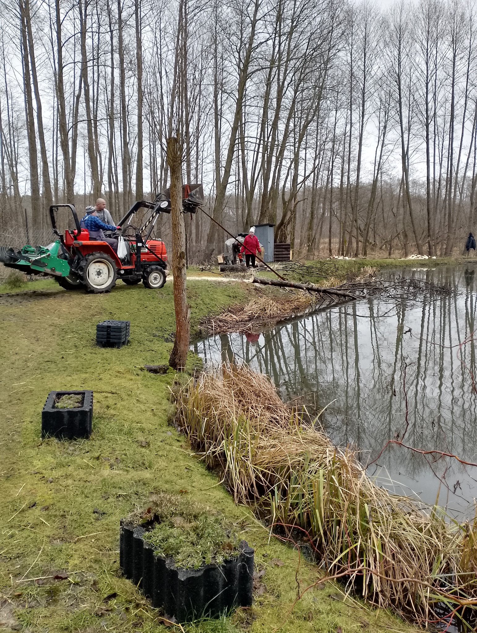 Prace porządkowe nad brzegiem stawu: traktor z kosiarką, ekipa usuwa gałęzie, czarne donice na brzegu, odbicie drzew w wodzie.