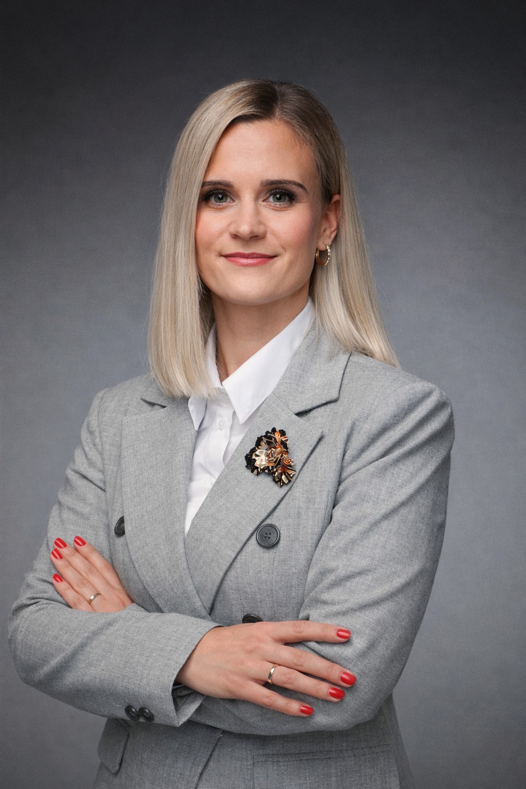 Portret kobiety w szarym garniturze z broszką, skrzyżowane ręce, blond włosy, na szarym tle. Profesjonalny wizerunek.