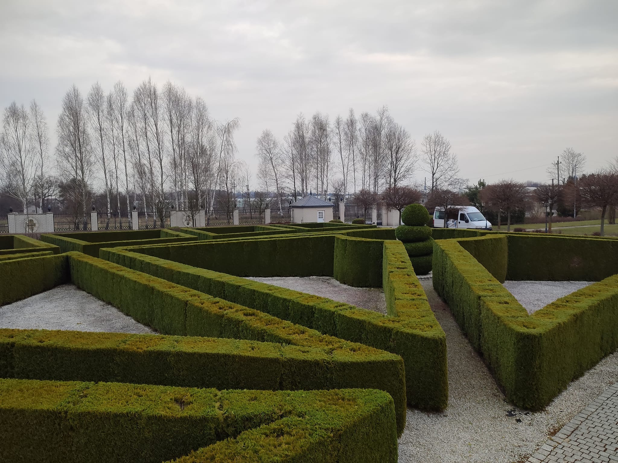 Geometryczny labirynt z żywopłotu bukszpanowego, wypełniony białym żwirem, z widocznym busem firmy ogrodniczej w tle, pod pochmurnym niebem.