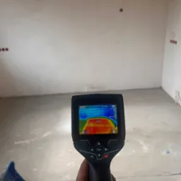 Pomiar temperatury ściany termowizją w surowym, otynkowanym pomieszczeniu z widocznymi puszkami elektrycznymi.