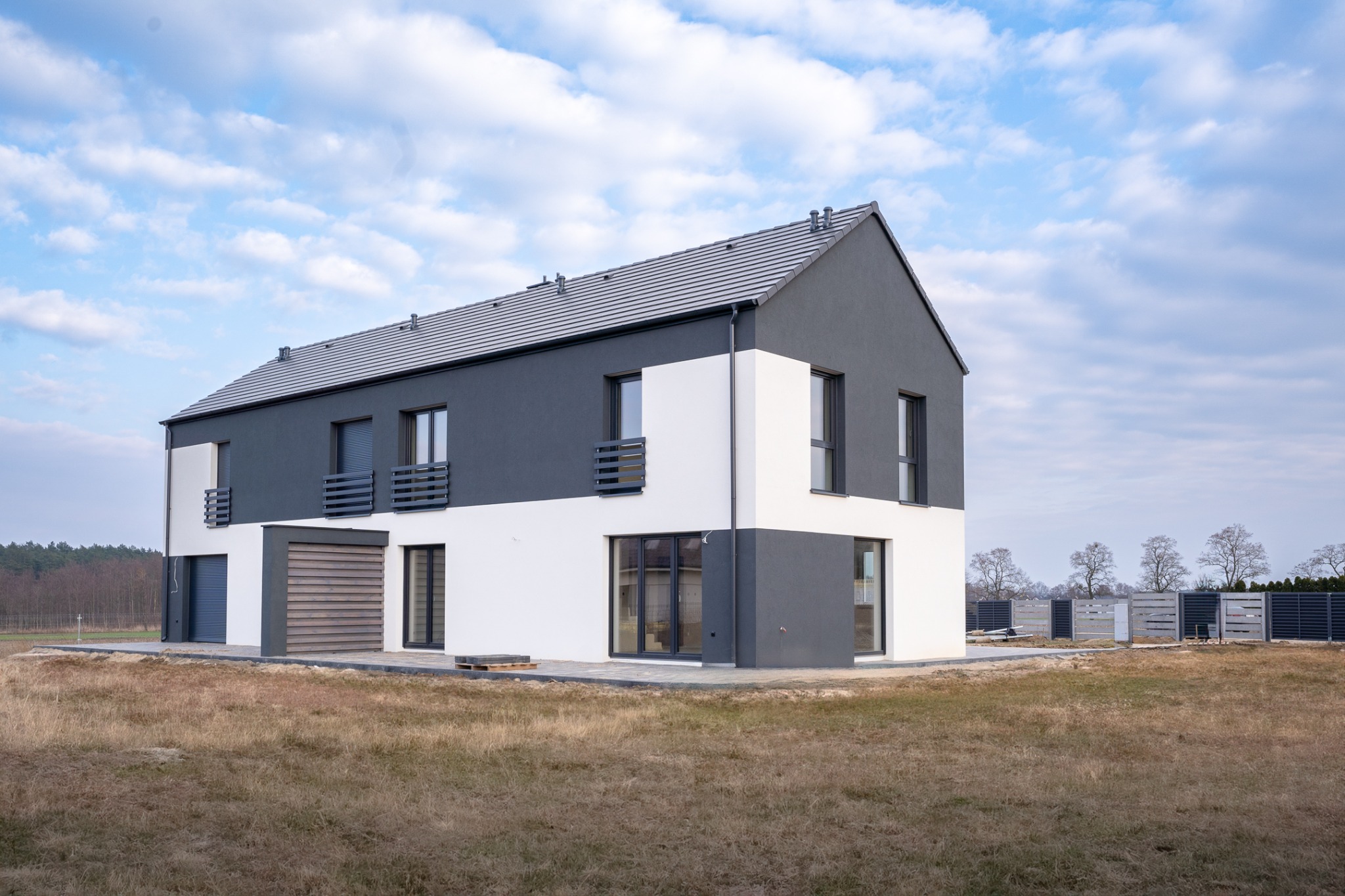 Nowoczesny, dwukondygnacyjny dom jednorodzinny z płaskim dachem i elewacją w kolorze białym i grafitowym, otoczony trawnikiem i ogrodzeniem panelowym.