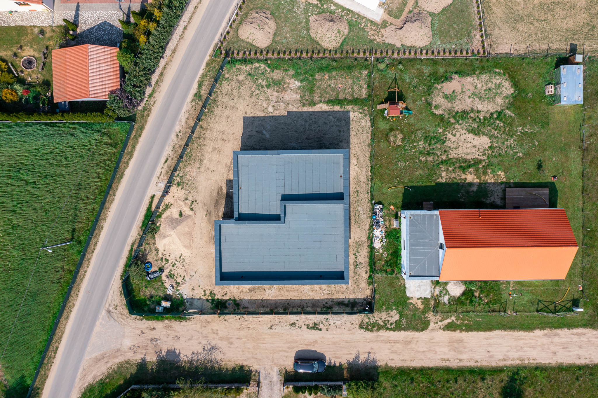 Widok z drona na budynek z szarym, płaskim dachem w trakcie budowy, otoczony niezagospodarowanym terenem, obok dom z czerwonym dachem, droga dojazdowa i zaparkowany samochód.