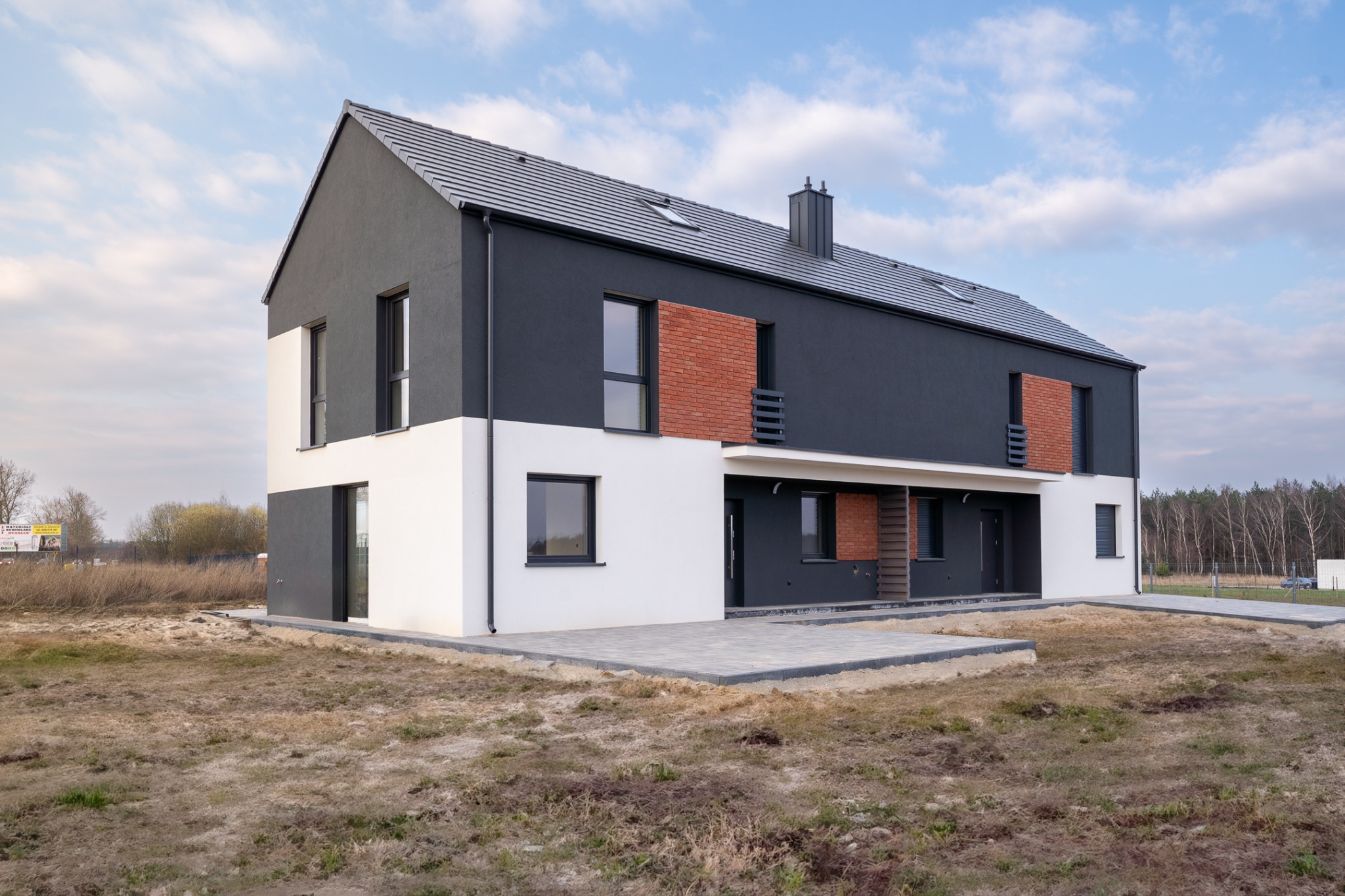 Nowoczesny, dwukondygnacyjny dom bliźniak z szarą elewacją, fragmentami cegły i białym parterem, widoczny z zewnątrz na tle błękitnego nieba.
