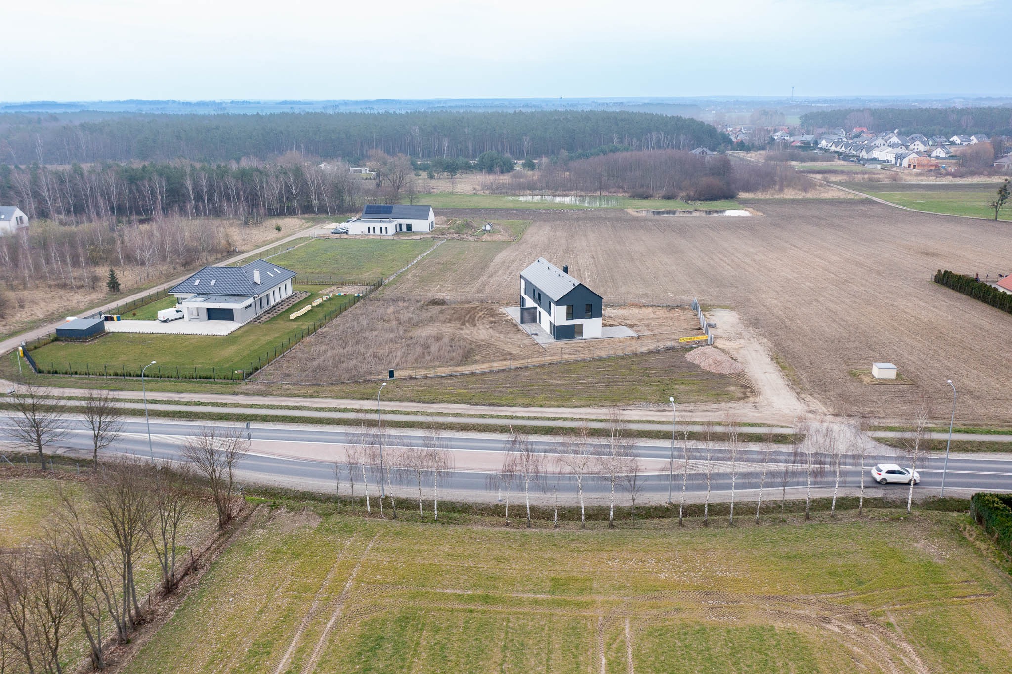 Szeroki kadr z lotu ptaka na trzy nowoczesne domy jednorodzinne na obrzeżach miasta, oddzielone polami i drogą z jadącym białym samochodem.