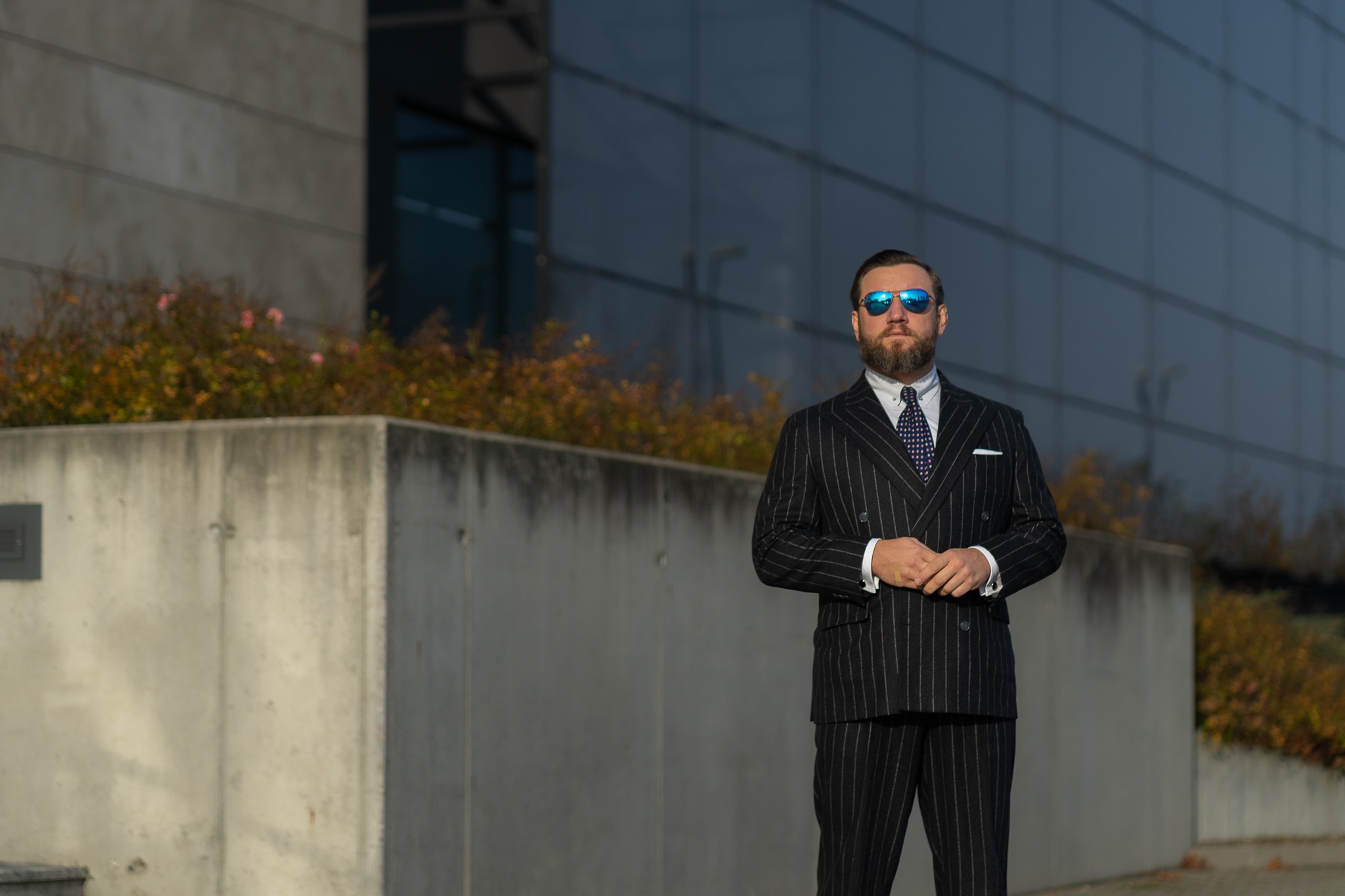 Mężczyzna w ciemnym garniturze w prążki i błękitnych okularach przeciwsłonecznych stoi na tle nowoczesnego budynku z przeszkloną fasadą.