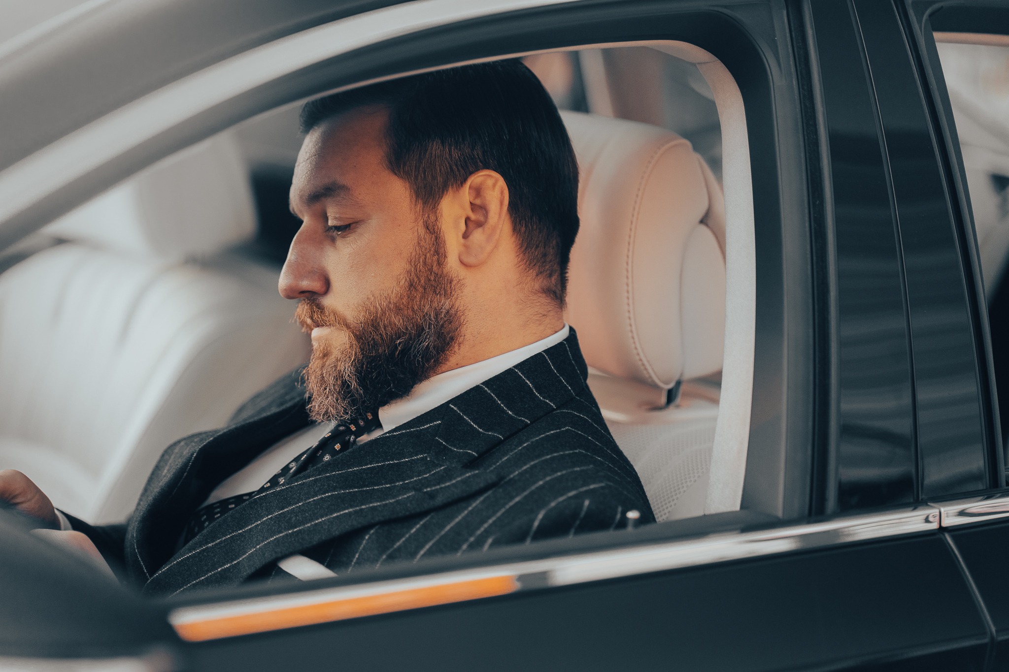 Mężczyzna z brodą w garniturze w paski, siedzący na jasnej skórzanej tapicerce luksusowego samochodu, z profilu, z zamkniętymi oczami.