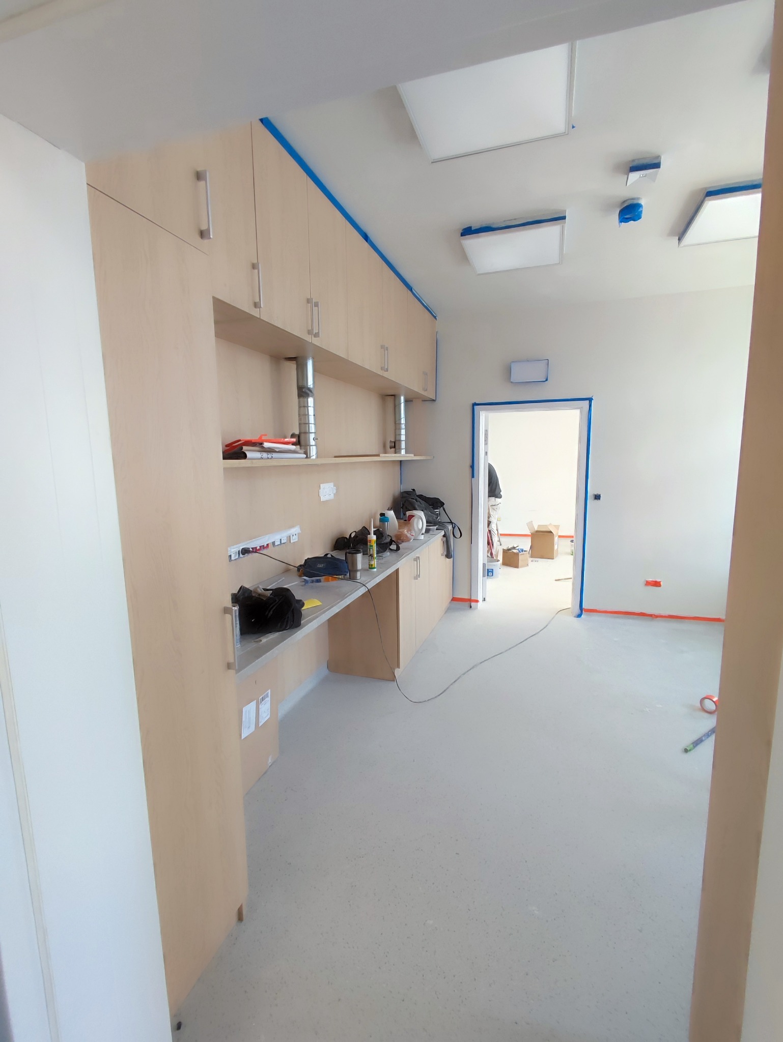 Przygotowania do prac wykończeniowych: zabezpieczone taśmą malarską krawędzie ścian, sufitu i ościeżnic w pomieszczeniu z zamontowanymi szafkami kuchennymi i blatem roboczym, widoczny pracownik...