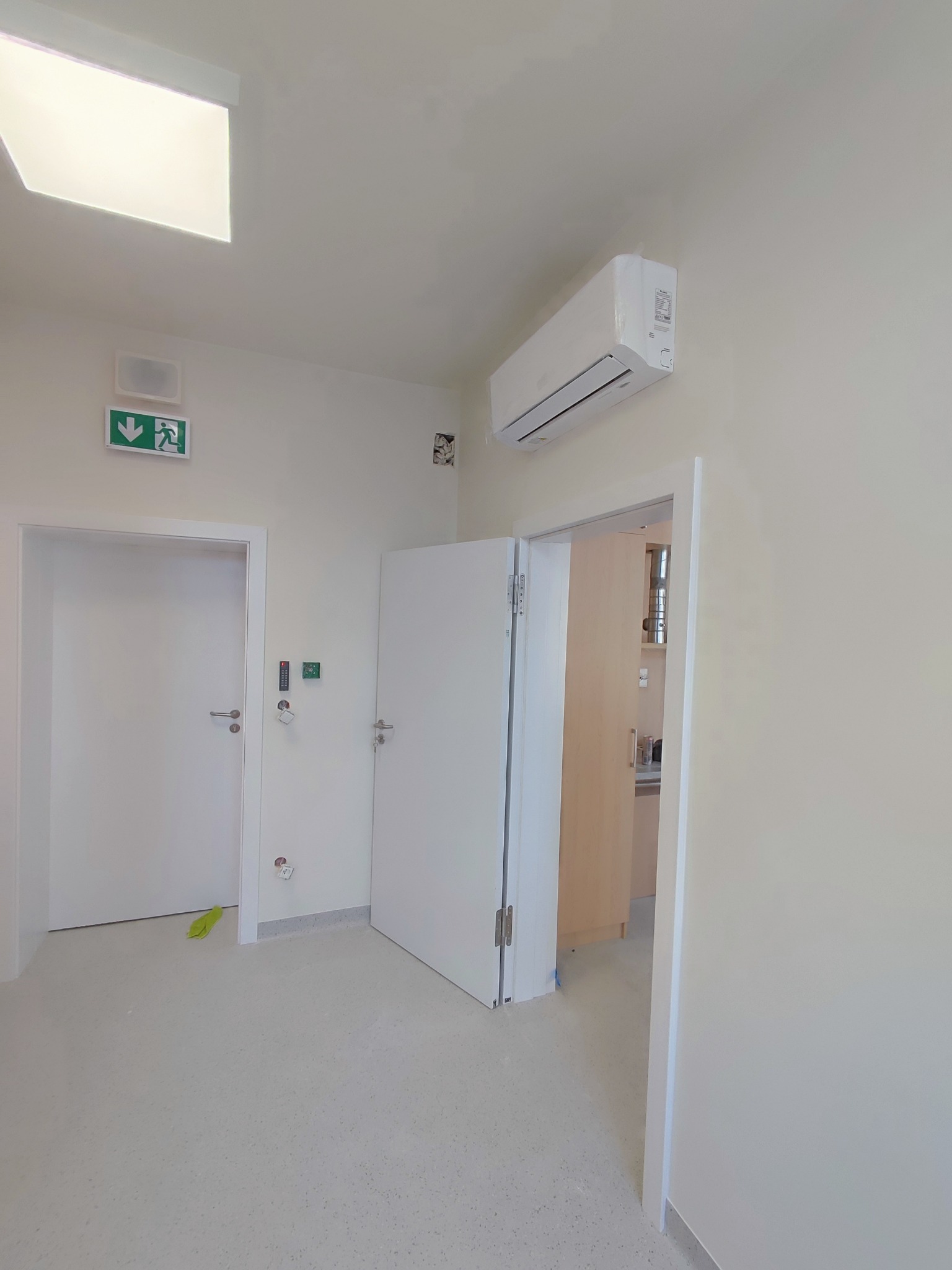 Wnętrze biura po remoncie, widoczne białe ściany, klimatyzator, dwoje białych drzwi, z czego jedne otwarte, fragment wyposażenia pomieszczenia za otwartymi drzwiami, na ścianie znak ewakuacyjny.