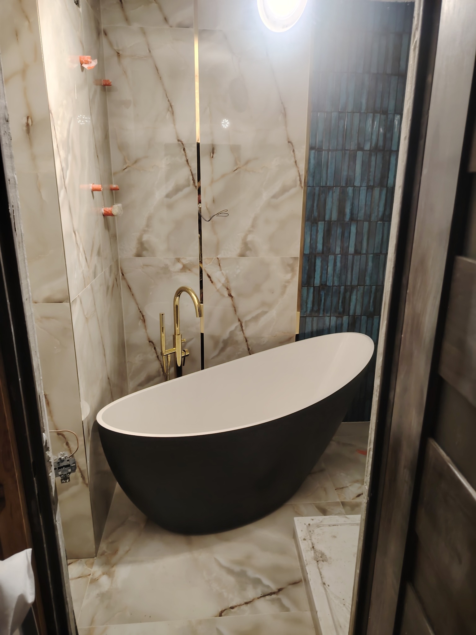 Nowoczesna łazienka z wolnostojącą wanną w kolorze czarnym, złotą armaturą i marmurowymi płytkami. Widoczne niebieskie płytki na ścianie i elementy instalacji.
