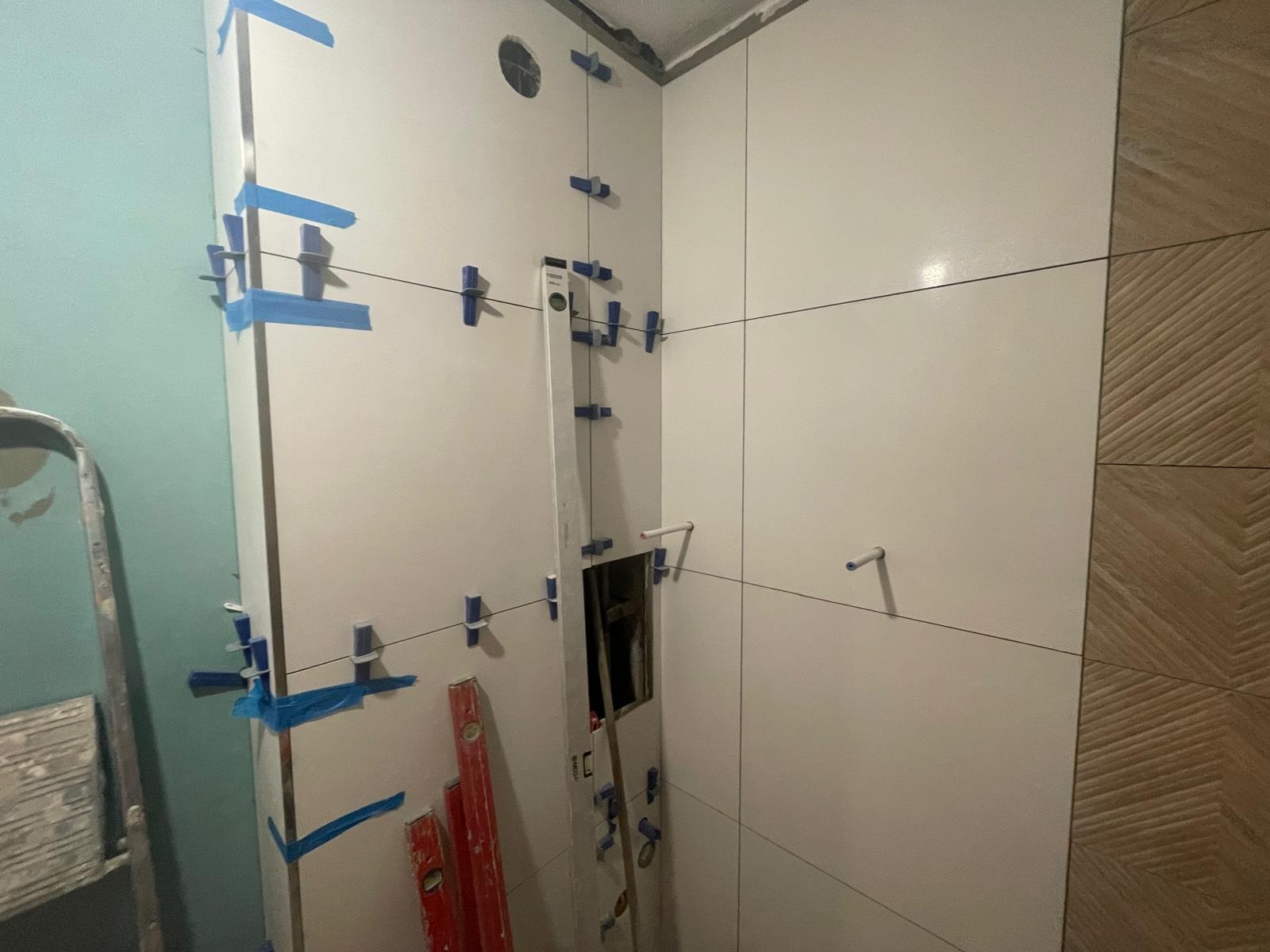 Białe płytki ścienne w łazience, w trakcie montażu, z użyciem poziomicy, sznurka i krzyżyków dystansowych. Fragment ściany z dekoracyjną, brązową płytką.