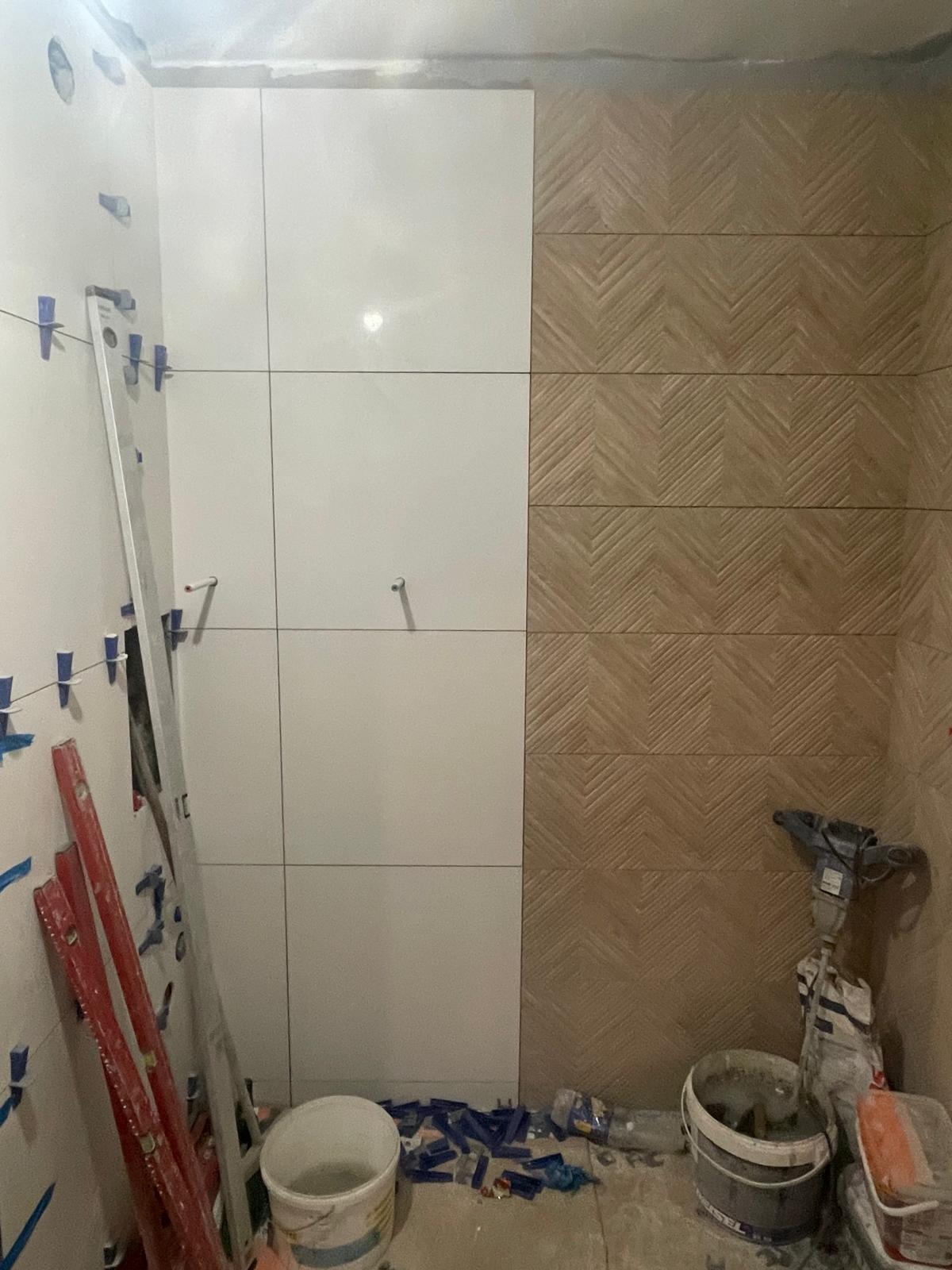 Układanie płytek w łazience: białe, kwadratowe płytki zestawione z dekoracyjnymi, drewnopodobnymi płytkami w jodełkę. Widoczne narzędzia glazurnicze i poziomice.