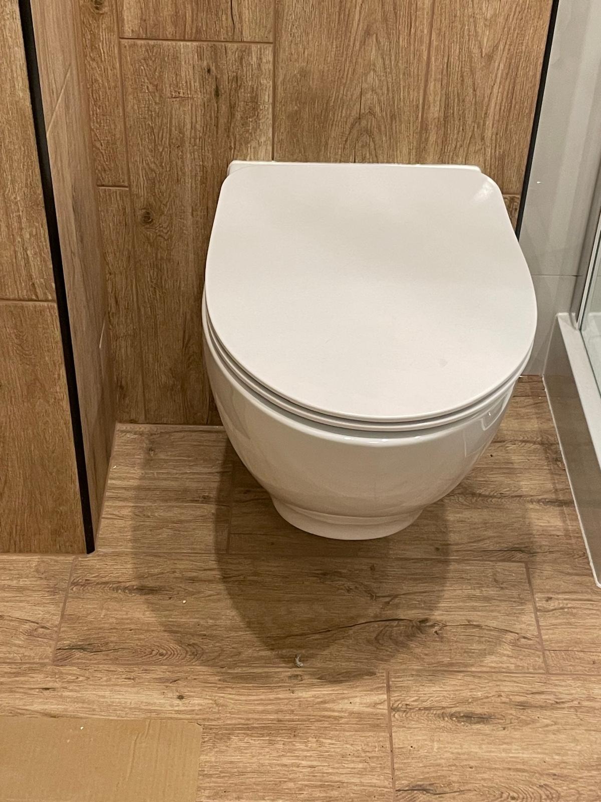 Nowoczesna toaleta wisząca w łazience z drewnopodobnymi płytkami na ścianach i podłodze. Widoczny fragment kabiny prysznicowej.