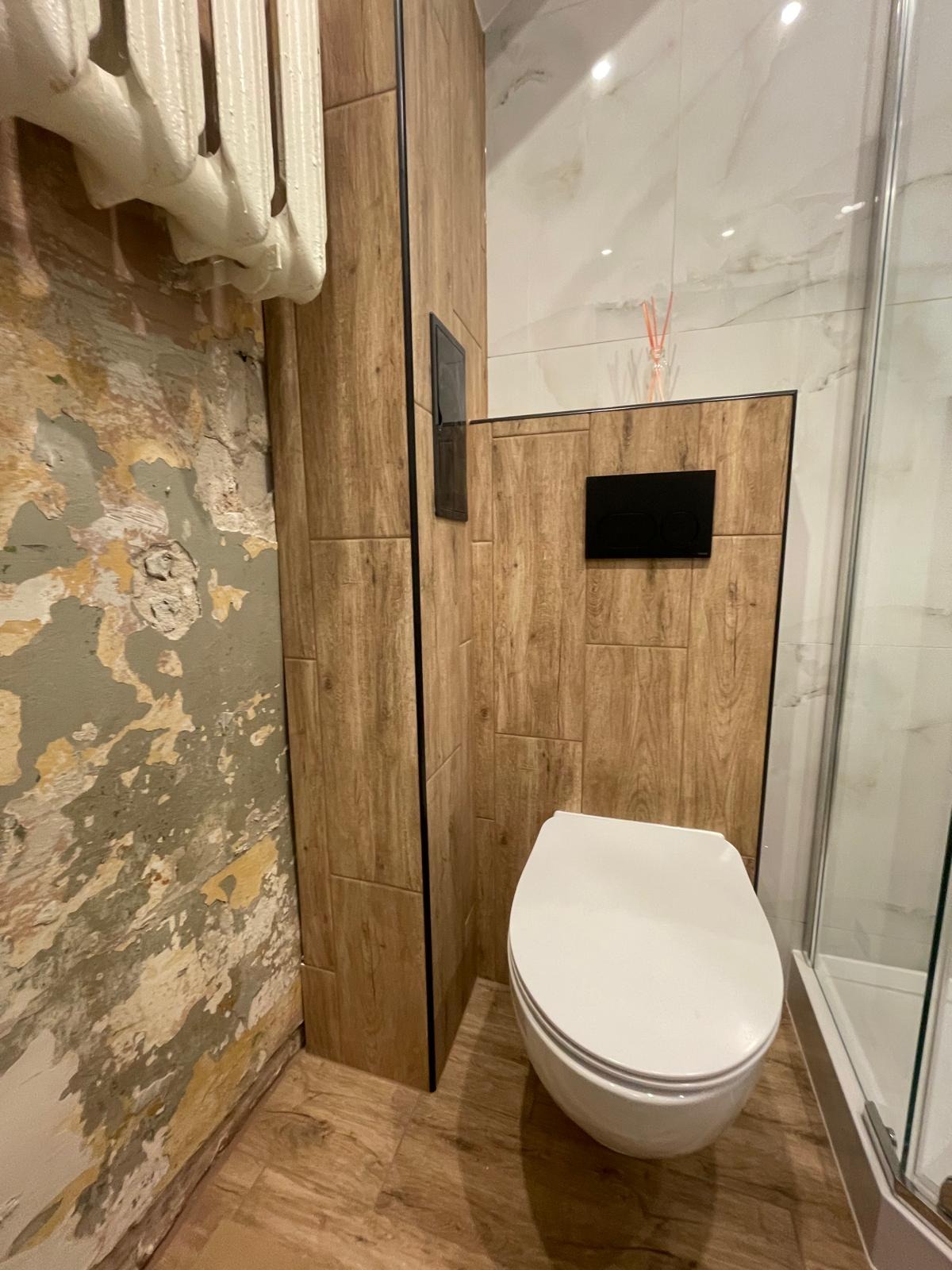 Łazienka w trakcie remontu: widoczna surowa ściana obok zabudowy WC z drewnopodobnych płytek. Fragment kabiny prysznicowej w tle.