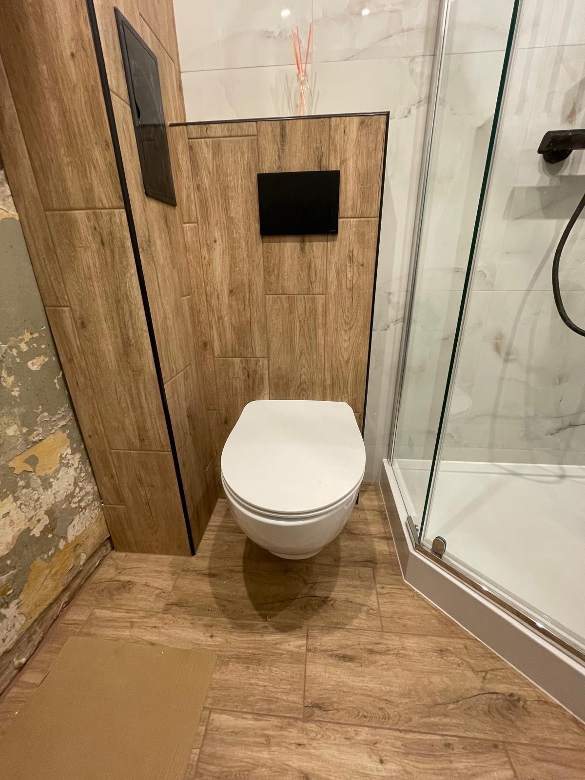 Nowoczesna toaleta wisząca w łazience z drewnopodobnymi płytkami i kabiną prysznicową. Widoczne fragmenty ściany przed wykończeniem.