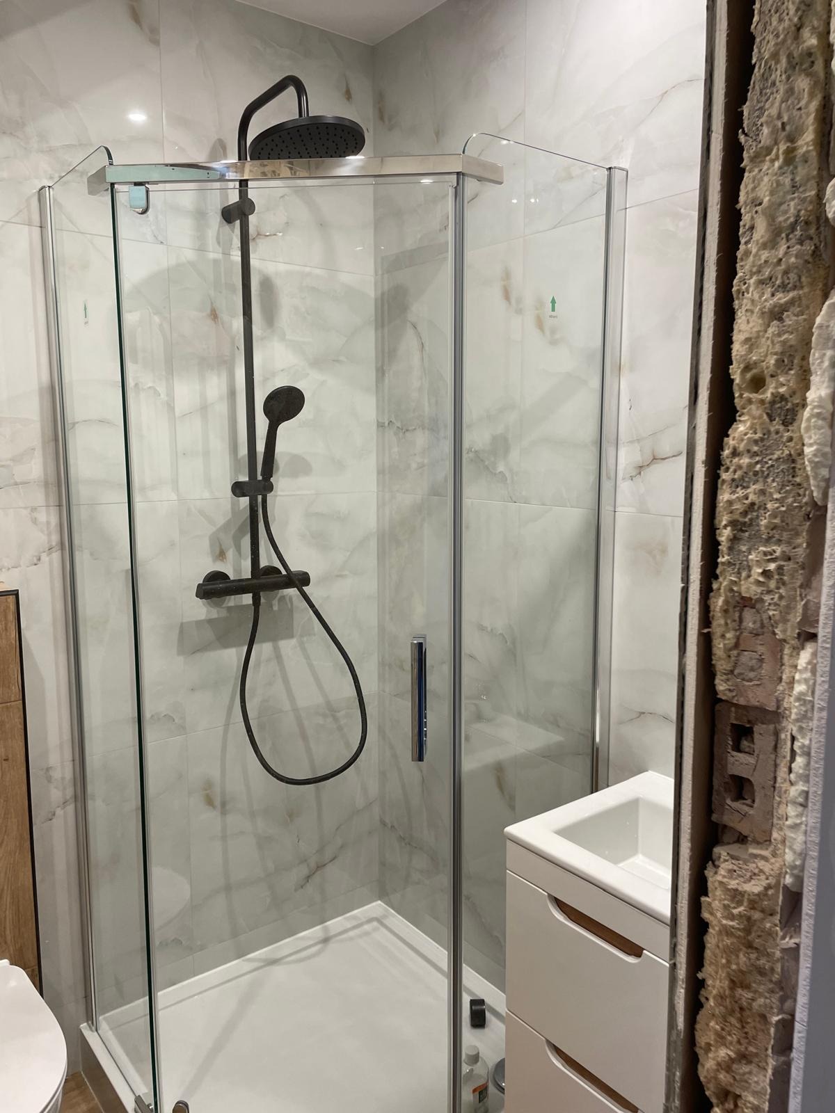 Nowoczesna kabina prysznicowa z deszczownicą i marmurowymi płytkami, obok fragment ściany w trakcie prac wykończeniowych. Widoczna umywalka z szafką.